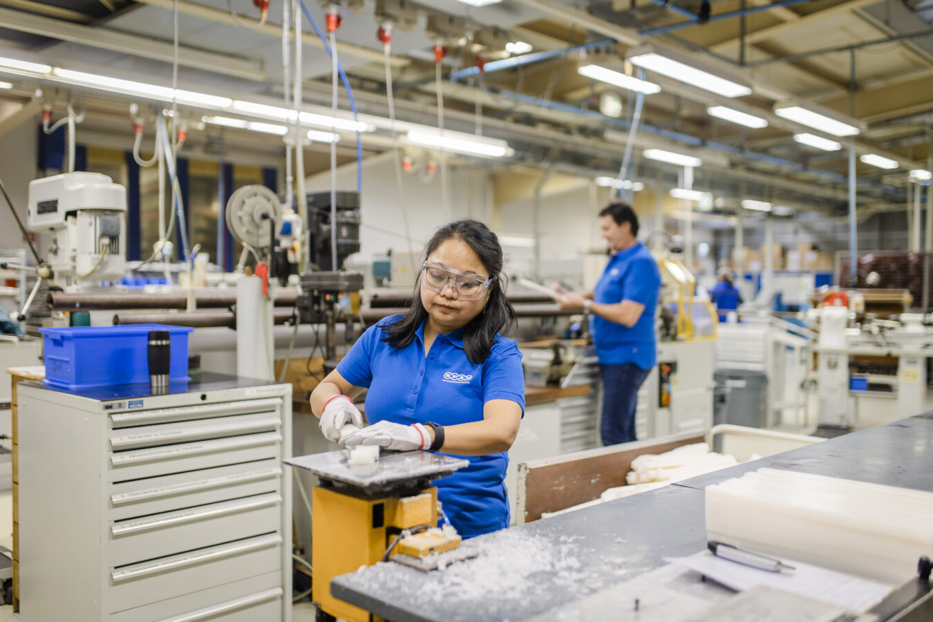 Eine Mitarbeiterin im Späh-Poloshirt bearbeitet mit Schutzbrille und Handschuhen in einer hellen Produktionshalle ein weißes Werkstück auf einer Maschine, während ein Späh-Kollege im Hintergrund arbeitet.