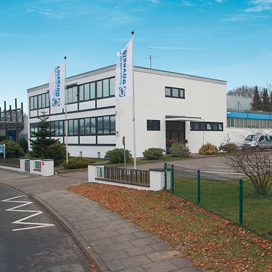 Das weiße, zweistöckige Bürogebäude der Firma Quadriga aus dem Jahr 1975 mit breiten Fensterfronten und zwei Fahnenmasten davor steht an einer Straße mit einem Zaun im Vordergrund.