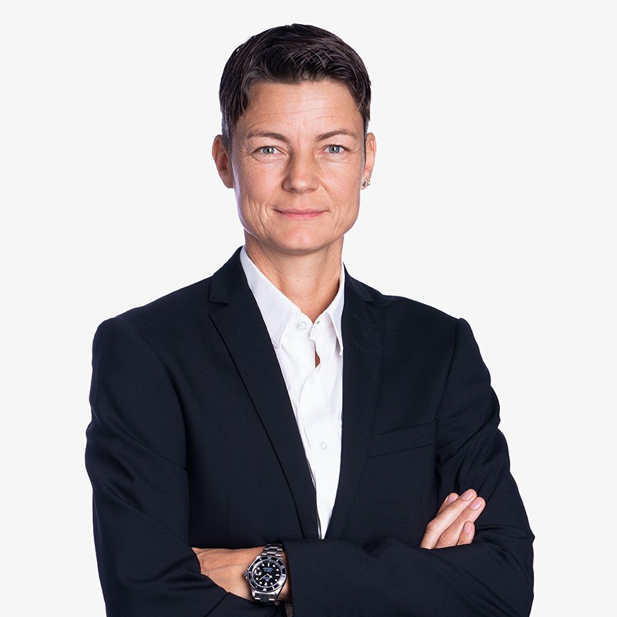 Das Business-Porträt zeigt Sandra Späh, eine Frau mit kurzen, dunklen Haaren und einem selbstbewussten Lächeln in die Kamera, die einen dunklen Anzug trägt und ihre Arme verschränkt hat.