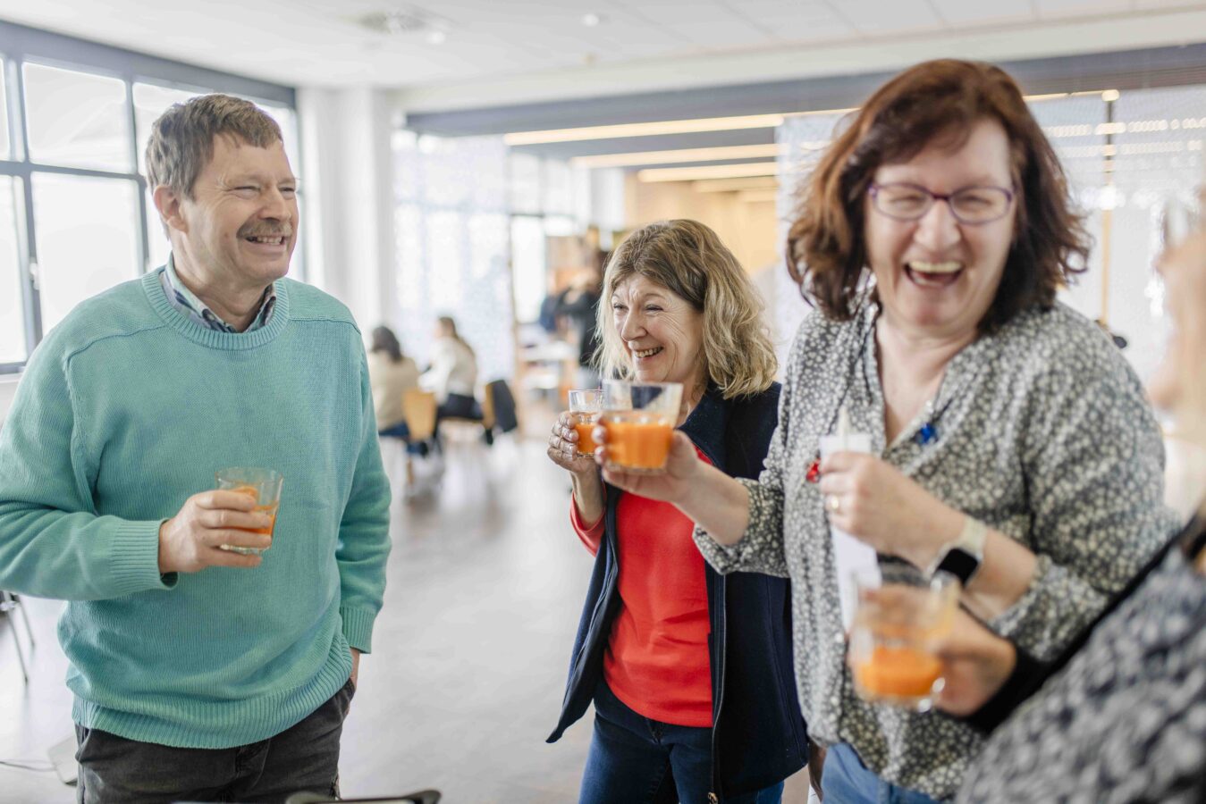 Eine Gruppe von Kollegen lacht und prostet sich mit Gläsern voll orangefarbenem Saft in einem hellen Gemeinschaftsraum zu.