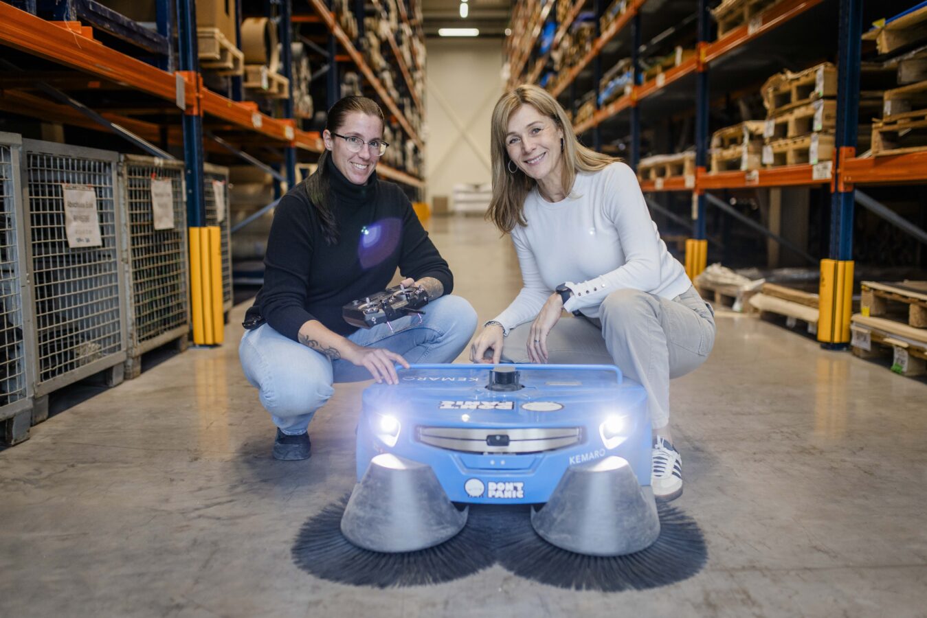 Zwei lächelnde Frauen knien in einem Lagergang mit hohen Regalen und Paletten neben einem kleinen, blauen, autonomen Kehrroboter der Marke KEMARO: Die eine Frau in dunkler Kleidung hält eine Fernbedienung, während die andere Frau in heller Kleidung auf den Roboter zeigt.