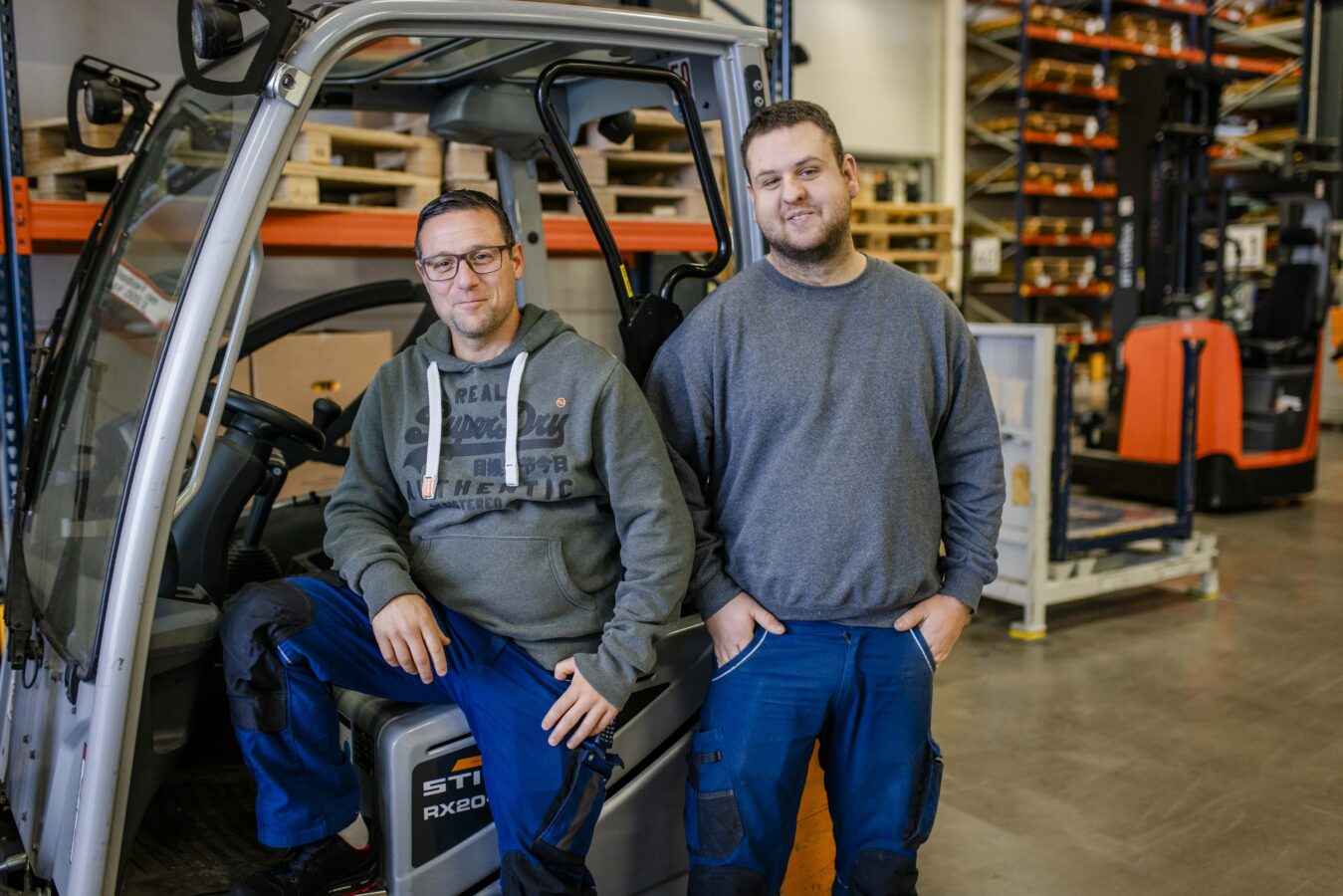 Zwei lächelnde Männer posieren in einer Lagerhalle der Späh-Gruppe, wobei der linke Mann in einem grauen Kapuzenpullover auf einem Gabelstapler sitzt und der rechte Mann in einem grauen Sweatshirt neben ihm steht.
