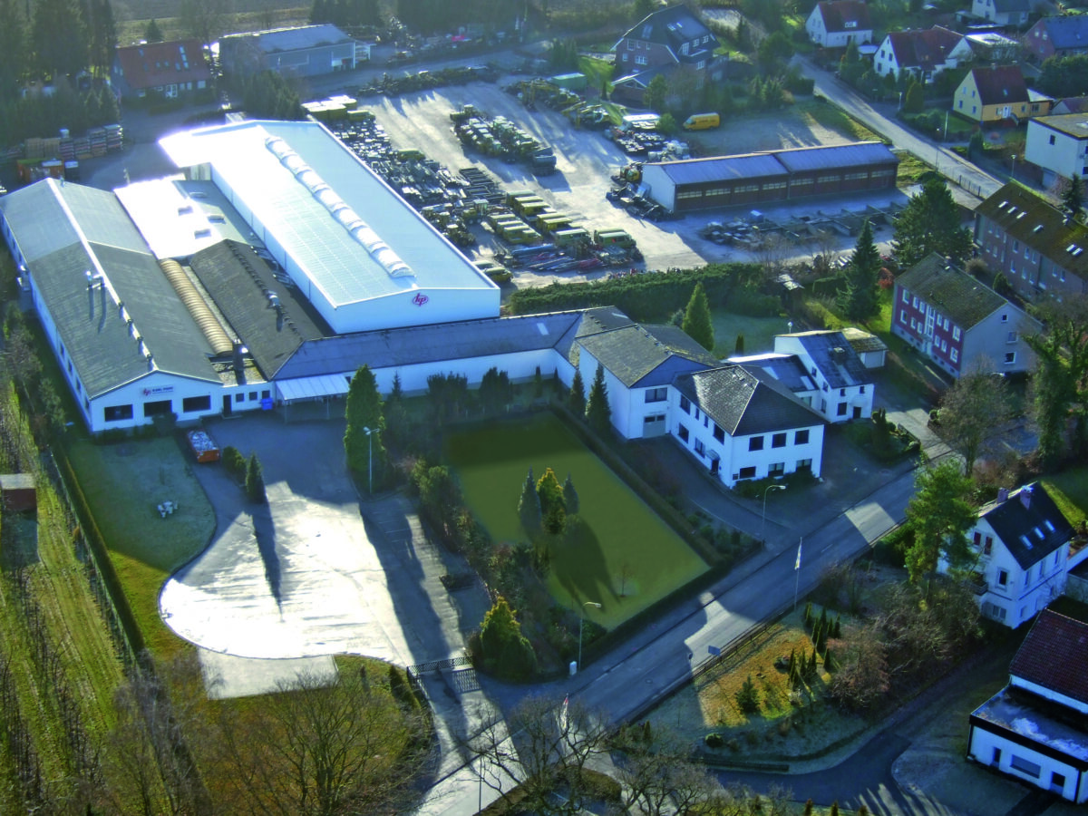 Eine Luftaufnahme zeigt das Firmengebäude der Karl Popp GmbH im Jahr 1972 mit einem langen Produktionshallen-Anbau und einem gepflegten Rasenbereich, umgeben von einem großen Lagerbereich mit Fahrzeugen und Material.