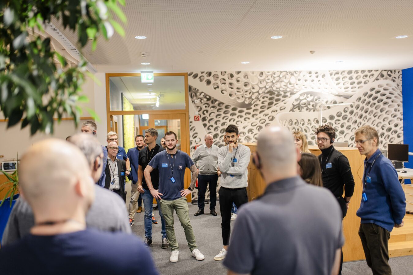 Eine Gruppe von Gästen hört aufmerksam zu, die vor einer auffälligen, weiß-gemusterten Wand in einem modernen Innenraum steht und eine Führung leitet.