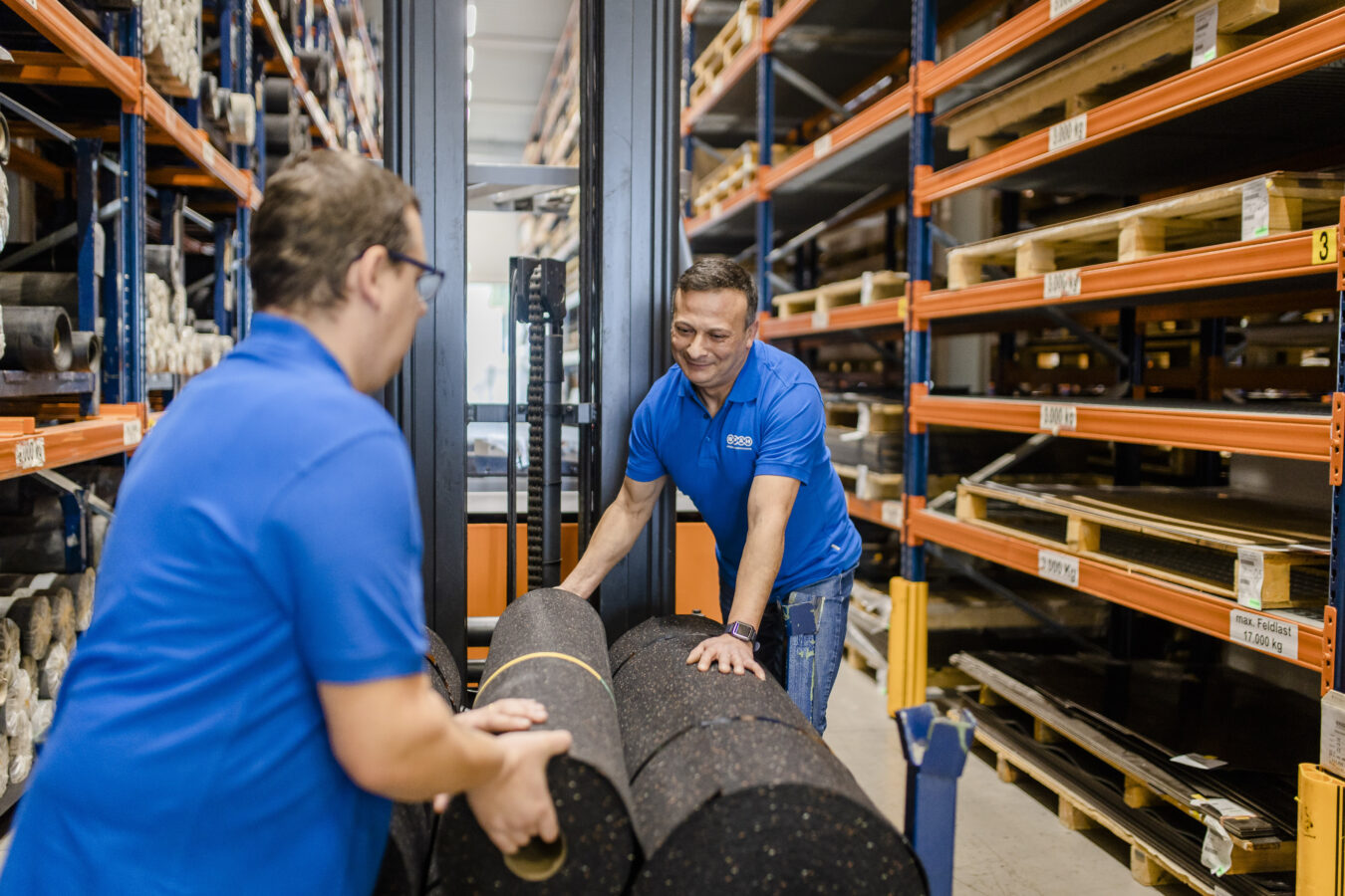 Zwei Mitarbeiter in blauen Späh-Poloshirts sind in einem Hochregallager dabei, große Rollen aus Elastomer-Material auf einem Flurförderzeug zu beladen.