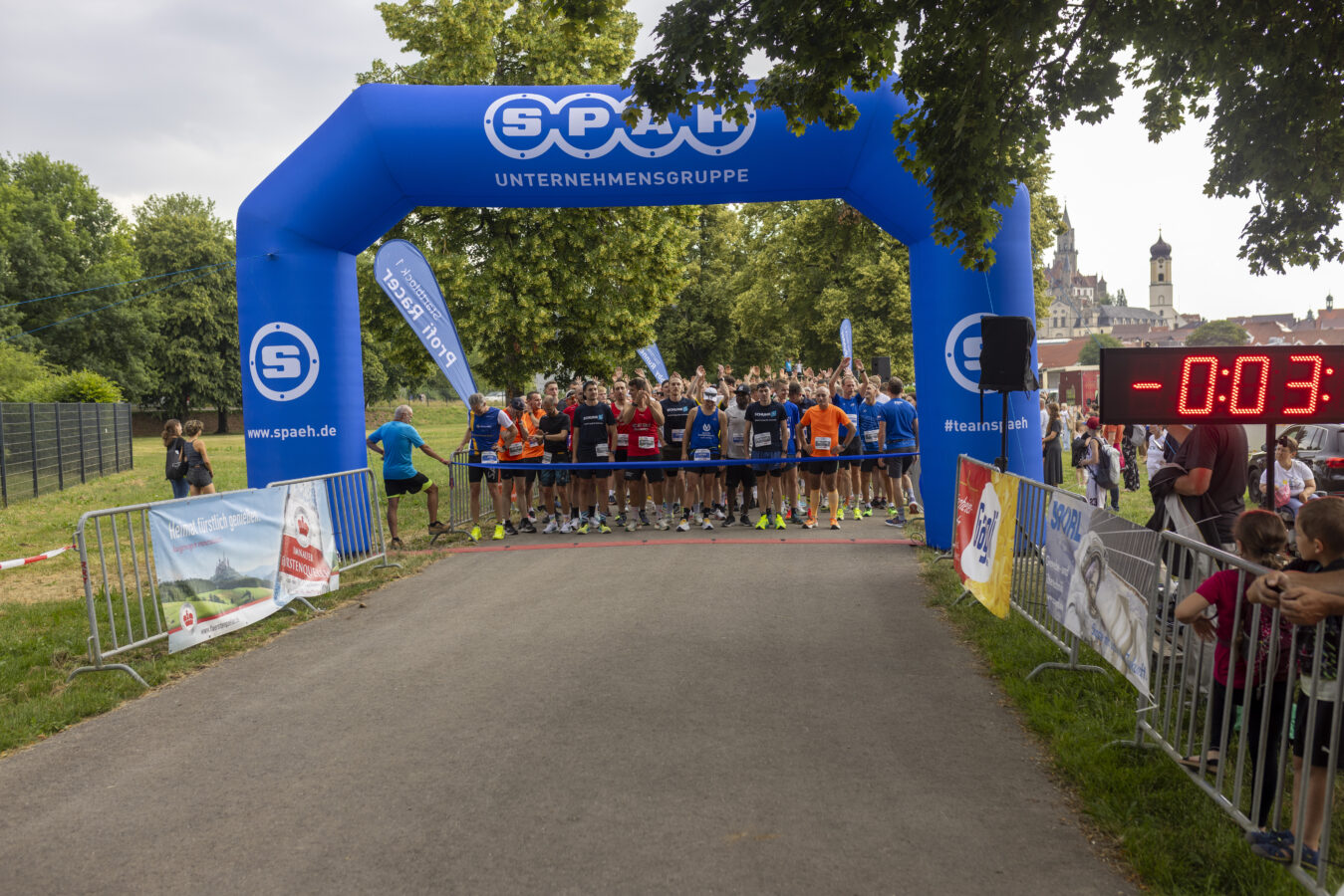 Eine große Gruppe von Läufern und Teilnehmern, viele davon in blauen, gelben oder orangefarbenen T-Shirts, steht vor einem großen Baum und hinter einem blauen SPÄH-Bogen, kurz vor dem Start eines Lauf-Events.
