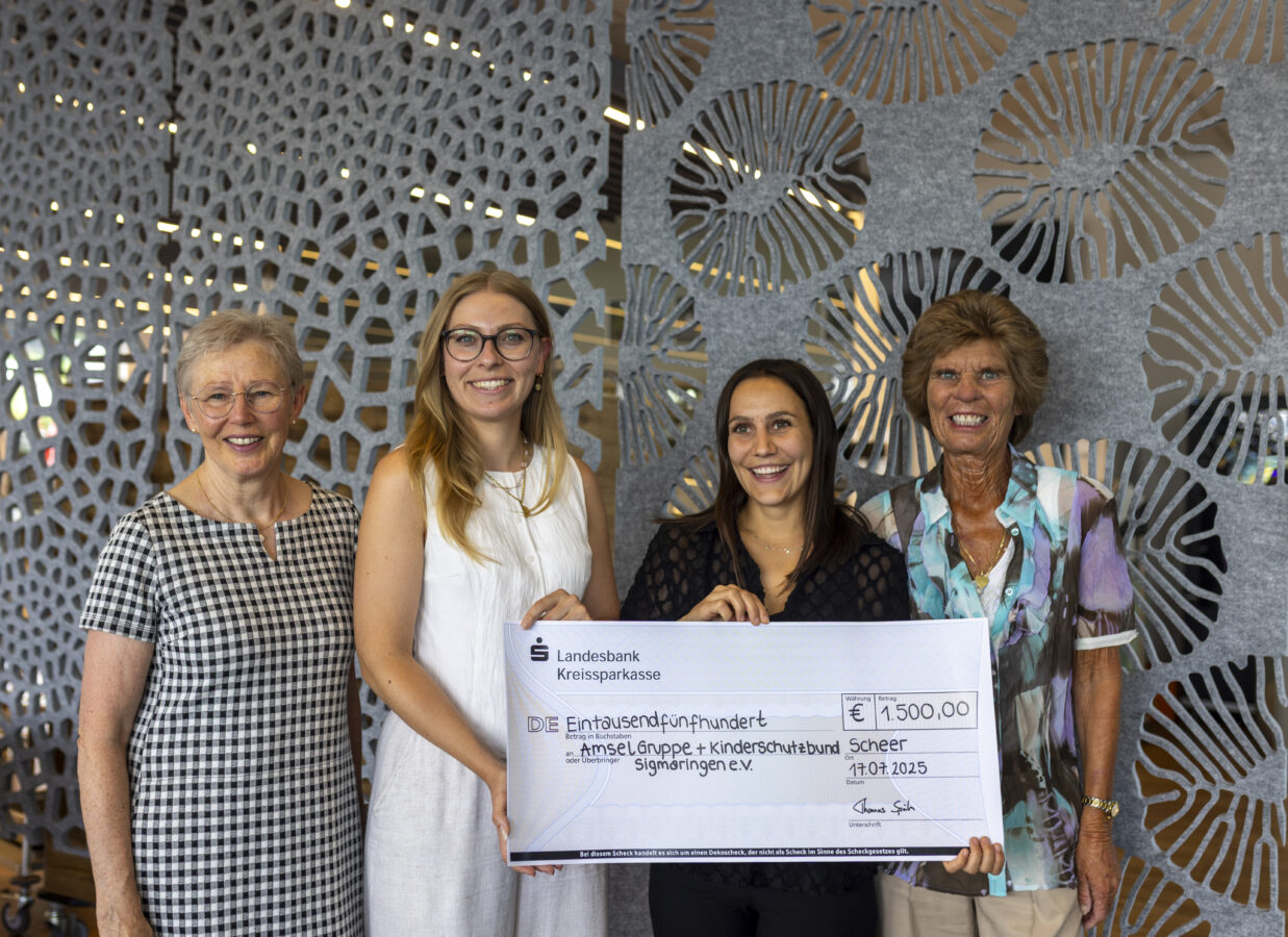 Vier Frauen stehen in einem modernen Innenraum vor einer grauen, gemusterten Trennwand und halten einen Scheck der Landesbank Kreissparkasse im Wert von € 1.500 für die Amselgruppe und den Kinderschutzbund Sigmaringen in die Kamera.