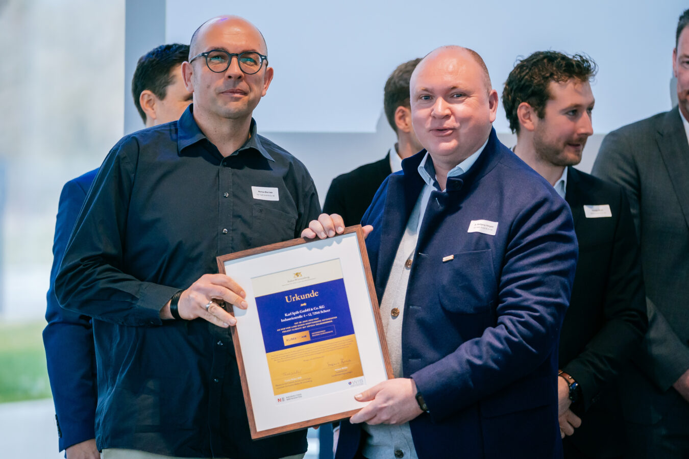 Zwei lächelnde Männer halten gemeinsam eine gerahmte Urkunde mit der Aufschrift Urkunde und dem Text "Wir haben ein klares Ja zu E-Mobilität und Co." vor dem Hintergrund einer Gruppe anderer Männer.