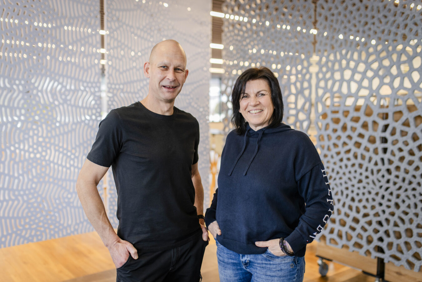 Das Bild zeigt eine lächelnde Frau in einem dunkelblauen Kapuzenpullover und Blue Jeans neben einem lächelnden, glatzköpfigen Mann in einem schwarzen T-Shirt, beide mit den Händen in den Taschen vor einer modernen Raumtrennwand mit einem grauen, wabenartigen Muster.