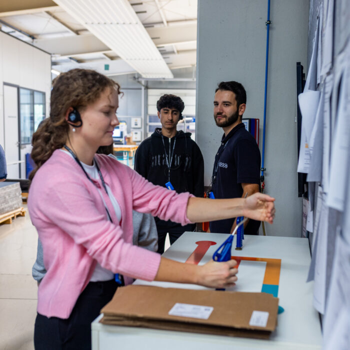 In der Produktionsabteilung befindet sich eine Gruppe von Auszubildenden. Eine Auszubildende testet eine Druckluftanwendung, während die anderen Auszubildenden den Vorgang beobachten.