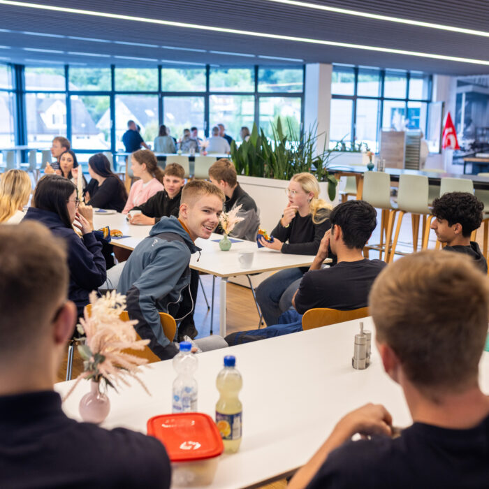 Alle Auszubildenden sitzen an einem Tisch in der Kantine um zu Frühstücken. Ein Auszubildender dreht sich lächelnd um und unterhält sich mit anderen Auszubildenden. 