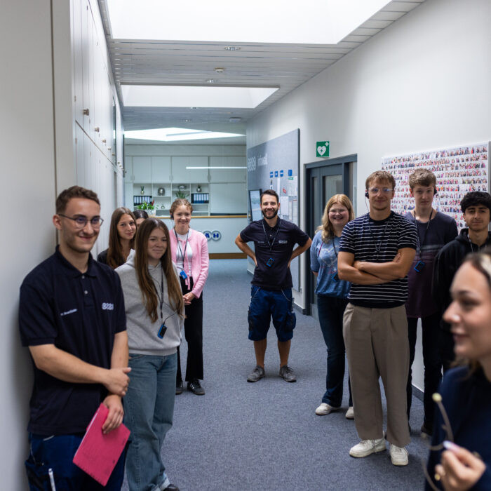 Eine Gruppe von Auszubildenden steht in einem Gang in der Verwaltung. Eine Auszubildende gibt den neuen Auszubildenden eine Führung durch das Unternehmen und erklärt ihnen etwas. 