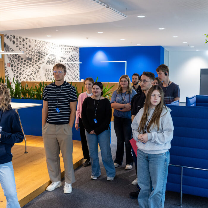 Eine Gruppe von Auszubildenden steht im Büro bei der QM, Buchhaltung und IT. Eine Auszubildende erzählt den neuen Auszubildenden etwas über die Abteilungen. 