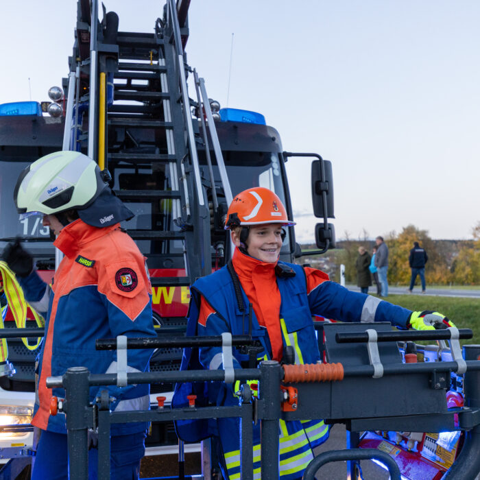 Zwei Mitarbeiter der Jugendfeuerwehr stehen im Korb der Drehleiter. Der jüngere Mitarbeiter lächelt. Der andere Mitarbeiter hält einen Wasserschlauch.