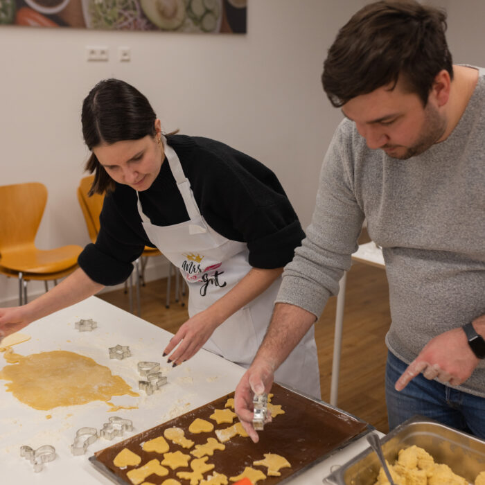 Zwei Späh-Mitarbeiter backen zusammen Weihnachtsgebäck in der Kantine. Es liegt ein ausgerollter Teig an der Seite mit vielen unterschiedlichen Ausstechern. Die ausgestochenen Plätzchen kommen auf ein Backblech.