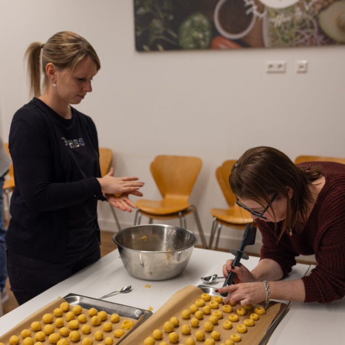 Zwei Späh Mitarbeiter backen zusammen Weihnachtsgebäck. Der eine Mitarbeiter rollt Bälle mit dem Teig und legt diese auf das Backblech. Der andere Mitarbeiter drückt kleine Vertiefungen in die Teigkugeln mit einem Werkzeug.