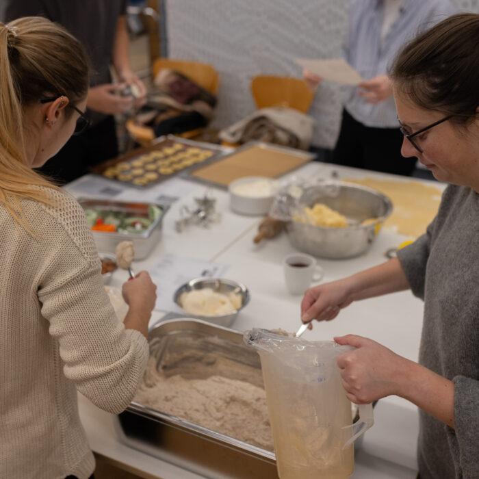 Zwei Späh-Mitarbeiter sind beim Backen von Weihnachtsgebäck. Vor ihnen steht eine große Schüssel mit Teig. Beide haben einen Löffel in ihren Händen. Im Hintergrund sind zwei Späh-Mitarbeiter mit einem Backblech voller Weihnachtsgebäck.
