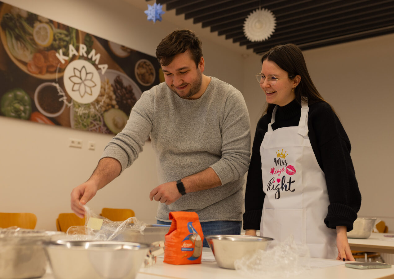 Zwei lächelnde Späh-Mitarbeiter in der Kantine beim Backen von Weihnachtsgebäck. Auf dem Tisch sind mehrere Schüsseln mit Zutaten.