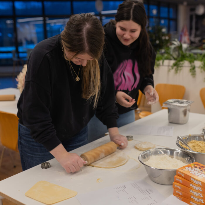 Zwei lächelnde Späh-Mitarbeiter rollen einen Teig aus um Plätzchen zu backen. Auf einem Backblech sind die ausgestochenen Plätzchen. In zwei unterschiedlichen Schüsseln befindet sich mehr Teig und Mehl.